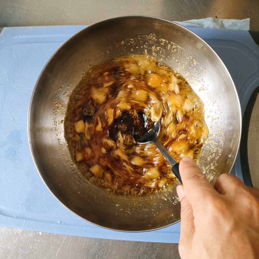 Une main mélangeant à la cuillère une marinade brun foncé contenant de lail haché et des oignons verts dans un bol en acier inoxydable.