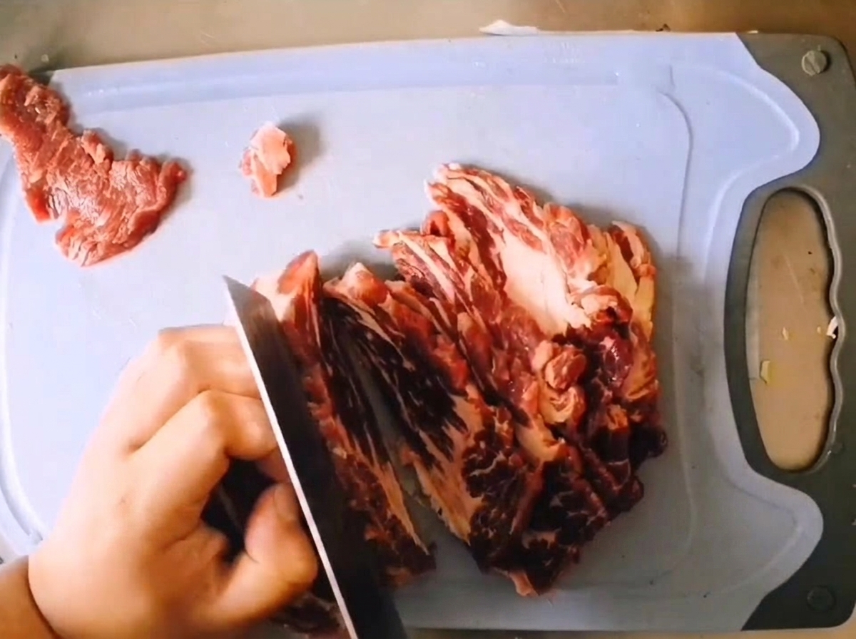 Découpe dun morceau de paleron de bœuf persillé en tranches épaisses avec un couteau bien aiguisé sur une planche bleu clair.