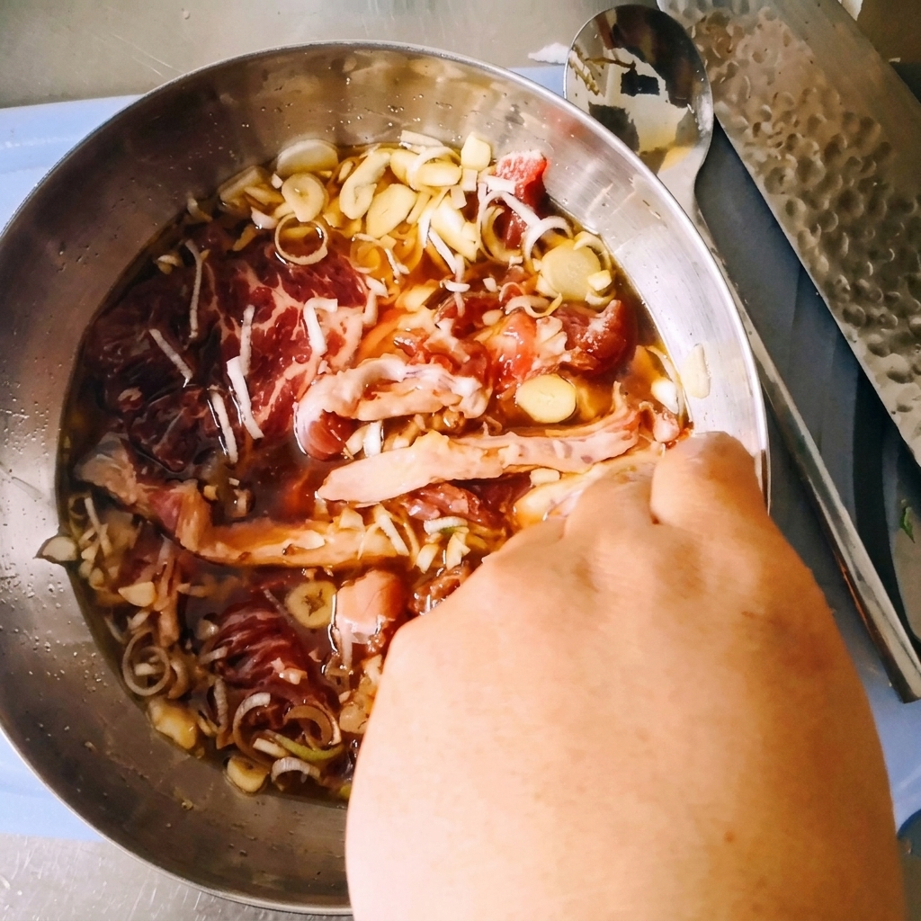 Une main mélangeant le paleron de bœuf cru tranché dans un bol en acier inoxydable avec la marinade au soja foncé, les oignons verts et lail.