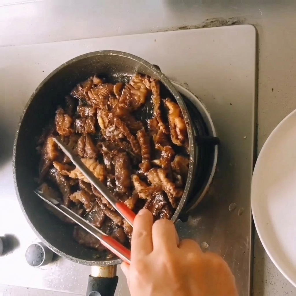 Morceaux de bœuf cuit mijotant dans une réduction de sauce riche et foncée à lintérieur dune poêle tout en étant remués avec une pince.