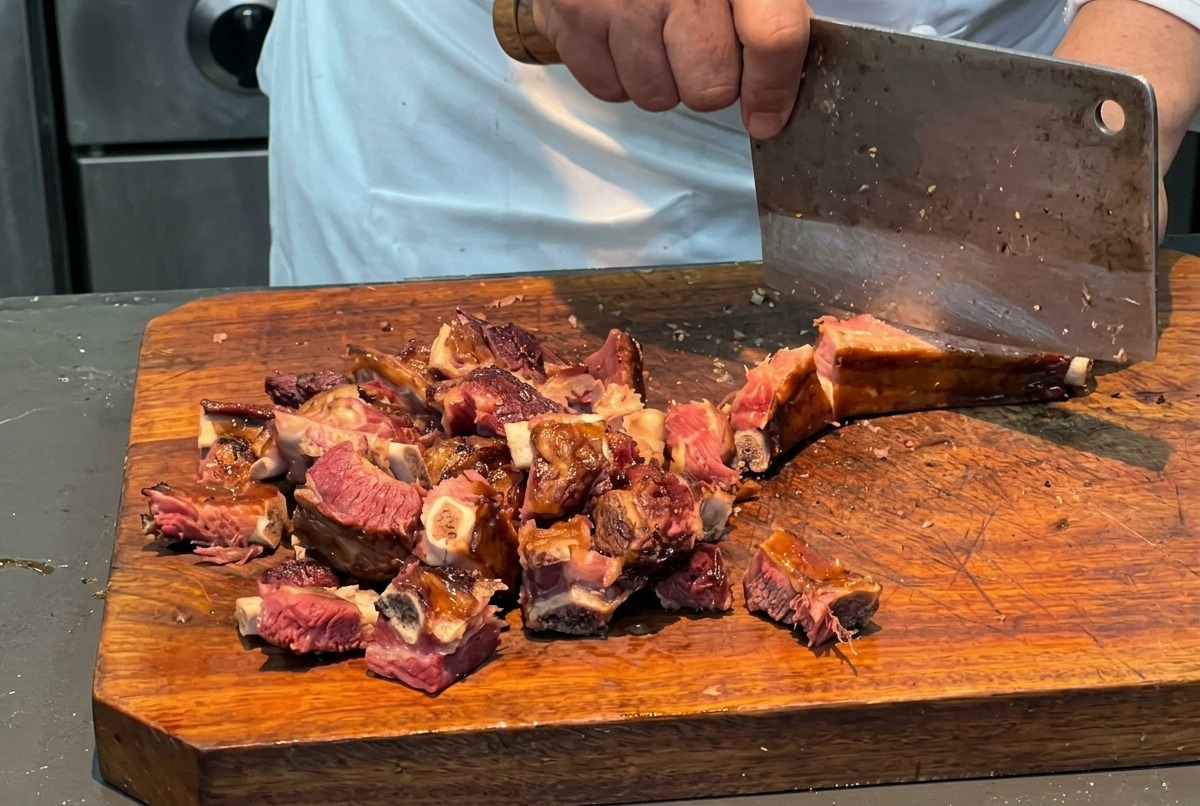 Un chef utilisant un grand couperet pour débiter un carré de porc fumé en morceaux réguliers sur une épaisse planche en bois.