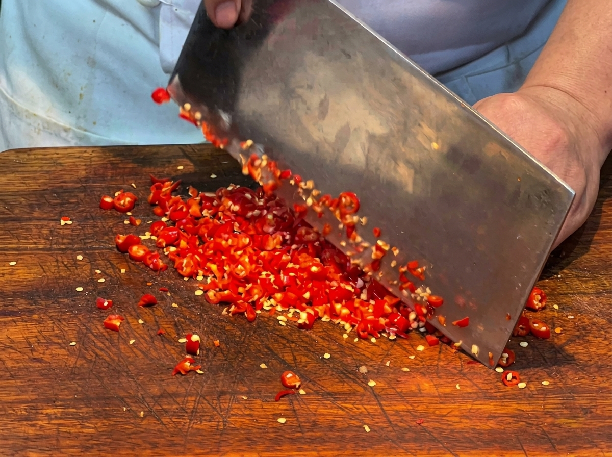 Gros plan sur des piments rouges frais en train dêtre finement hachés au couperet sur une planche en bois.