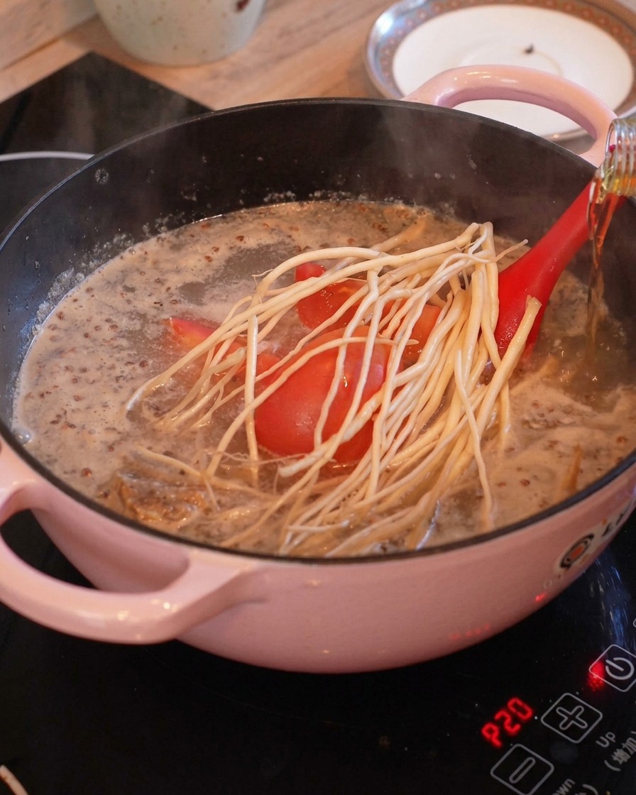 Des morceaux de tomates et de longues herbes à racines blanches dans une marmite bouillante avec un bouillon clair, pendant que du vin de cuisson est versé depuis une bouteille.