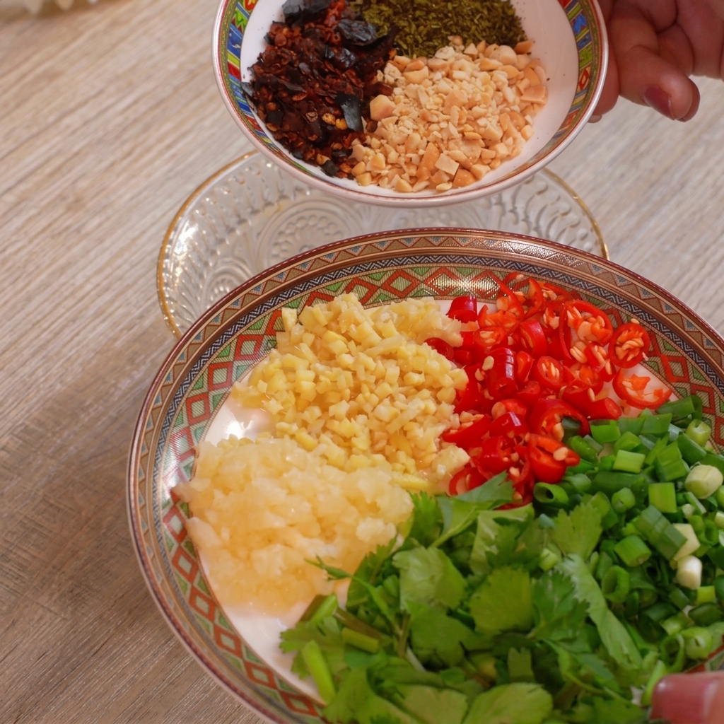Une assiette avec des petits tas séparés dail haché, de gingembre haché, de piments rouges coupés, doignons verts et de coriandre, avec un autre bol de cacahuètes et de flocons de piment tenu au-dessus.