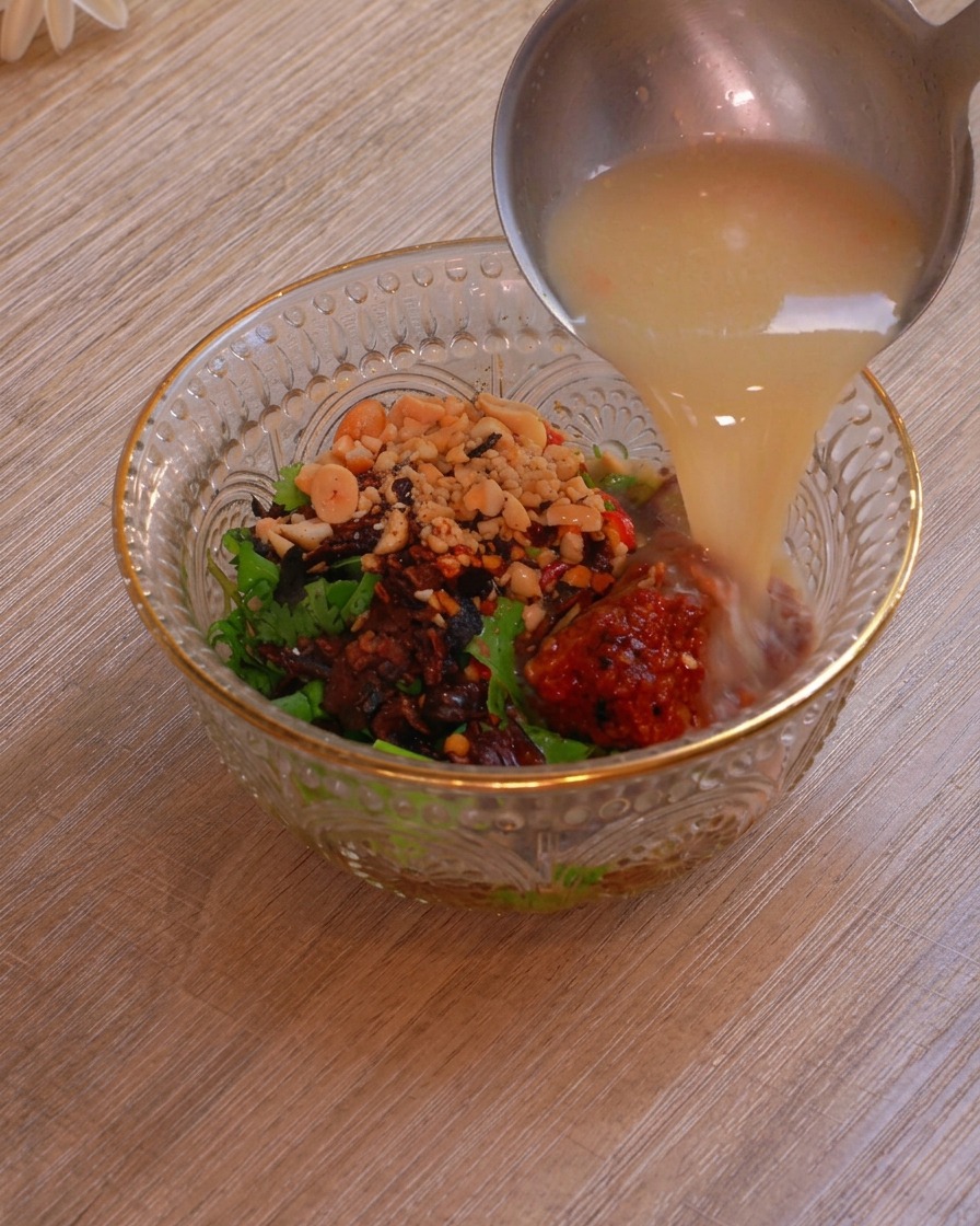 Une louche en métal verse un riche bouillon chaud et laiteux directement dans un bol en verre contenant des herbes, des cacahuètes, des flocons de piment et du tofu fermenté.