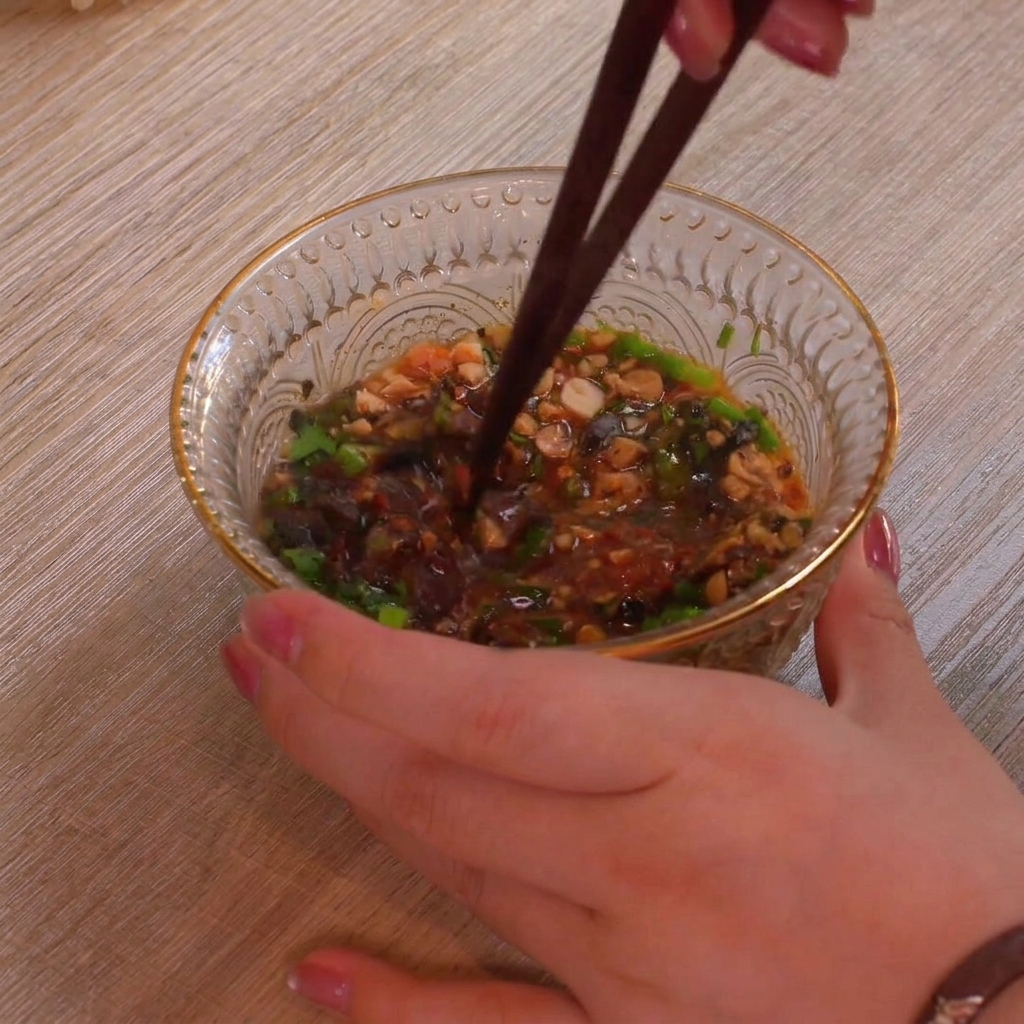 Un gros plan dun bol en verre transparent texturé rempli dingrédients colorés pour la trempette comme des oignons verts, des cacahuètes écrasées et des flocons de piment rouge, le tout mélangé soigneusement avec une paire de baguettes foncées.