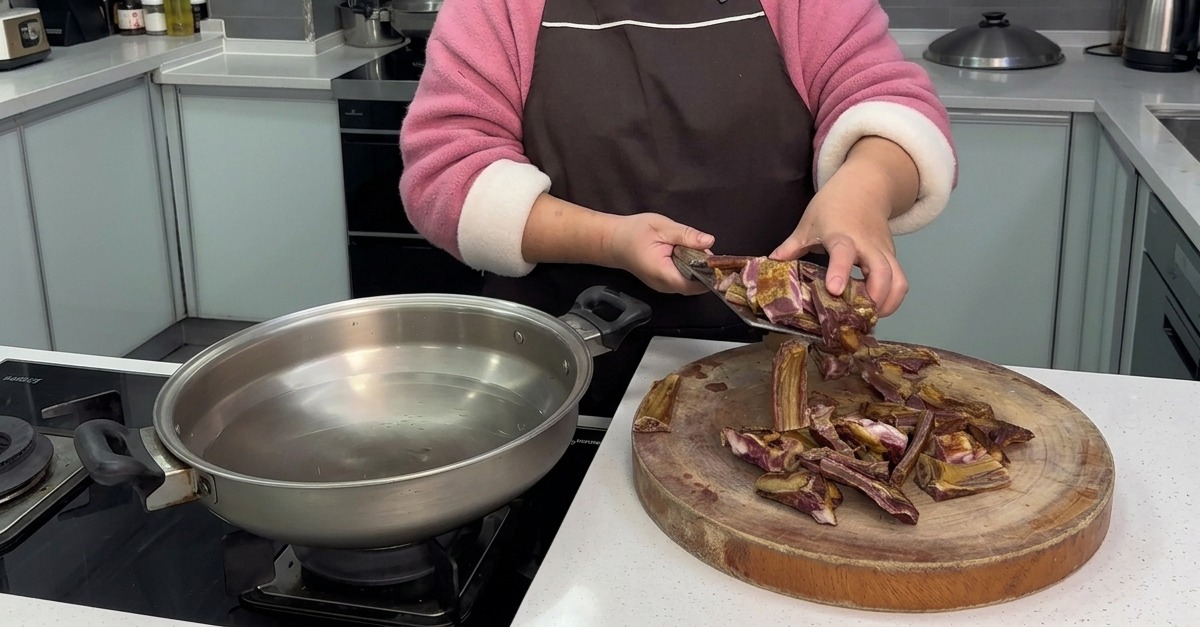 Une personne transférant des morceaux de travers de porc fraîchement coupés depuis une planche en bois vers une poêle en inox remplie deau froide.