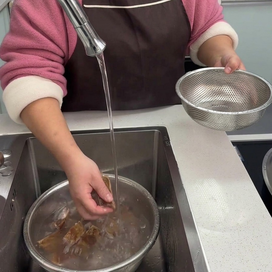 Des mains lavant soigneusement des morceaux de travers de porc blanchis sous un filet deau claire dans lévier de la cuisine.