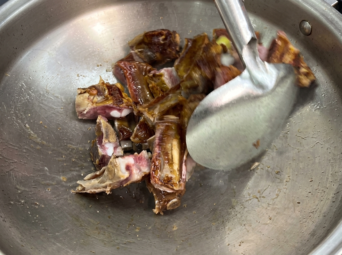 Une spatule métallique faisant sauter des morceaux de travers de porc et des tranches de gingembre frais dans un grand wok en inox.