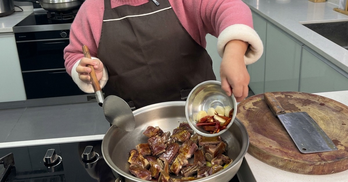 Une personne versant des gousses dail entières et des piments rouges séchés depuis un petit bol en métal dans un wok contenant le porc sauté.