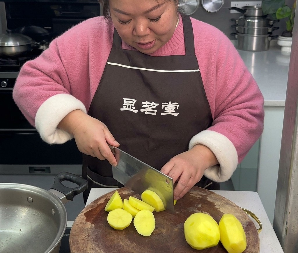 Une femme tablier marron coupant des pommes de terre jaunes pelées en morceaux biseautés sur une planche à découper en bois.