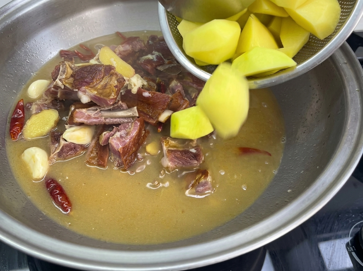 Des morceaux de pommes de terre dorées versés depuis une passoire dans une grande sauteuse contenant le bouillon frémissant, le porc et les piments.