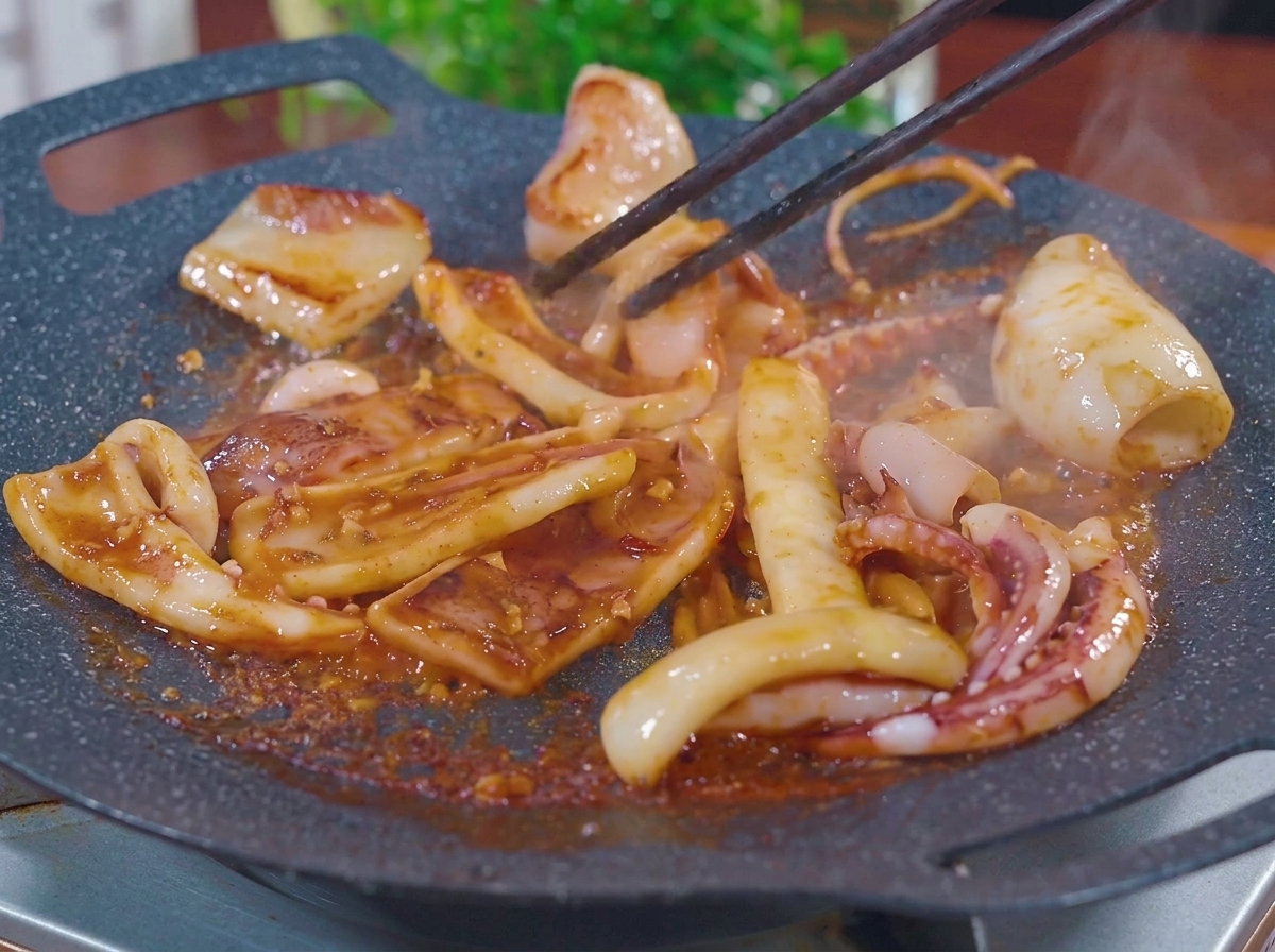 Lanières de calamar sautées avec des baguettes sur une plaque chaude, crépitant dans une sauce barbecue épaisse avec de la vapeur.