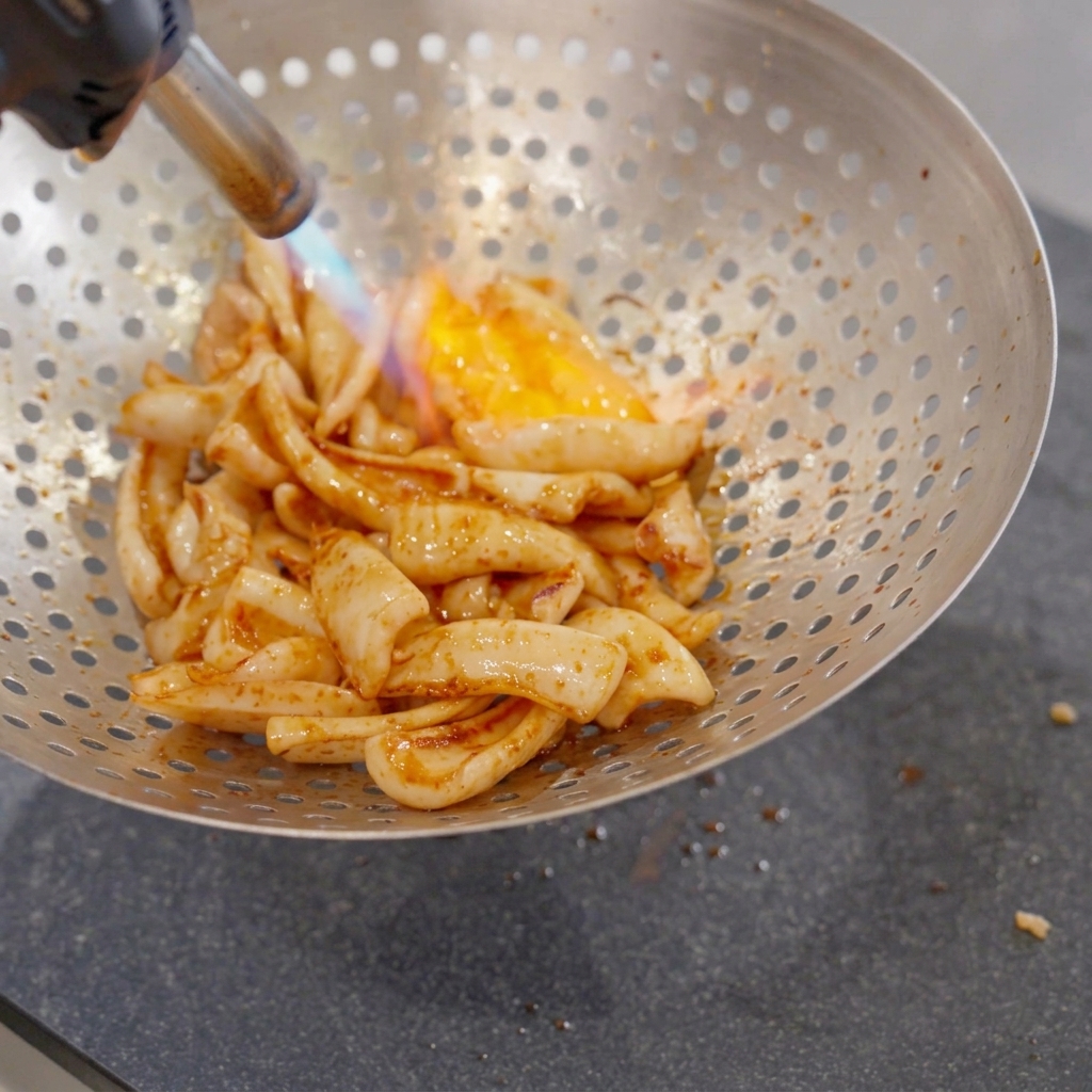 Des morceaux de calamar frit placés dans une grande écumoire métallique percée, saisis par la flamme bleue et orange dun chalumeau de cuisine.
