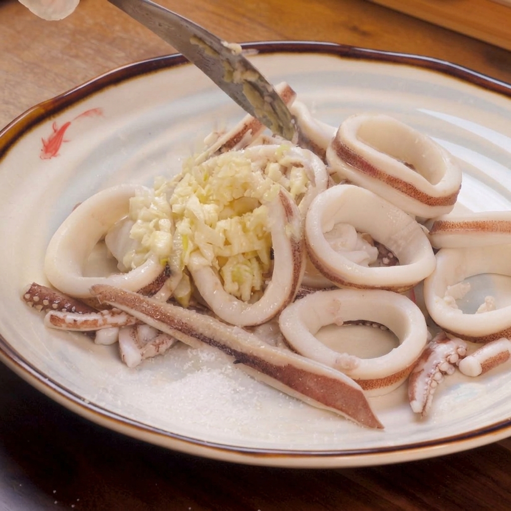 Anneaux de calmar blanchis disposés sur une assiette, surmontés dune bonne quantité dail haché avec une pincée de sel et de sucre sur le côté.