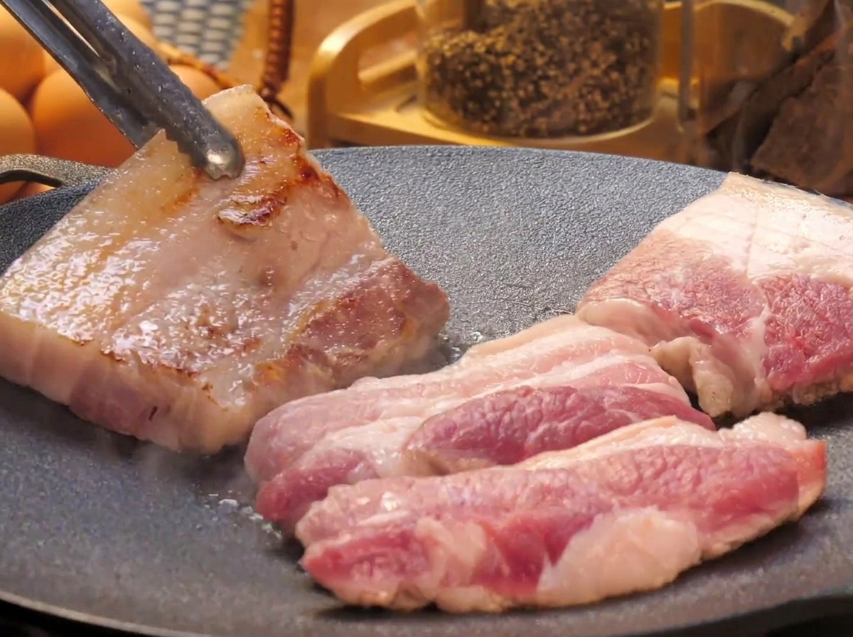 Tranches épaisses de poitrine de porc cuisant sur une plaque teppanyaki plate, une pièce étant retournée avec des pinces métalliques pour révéler sa surface dorée.
