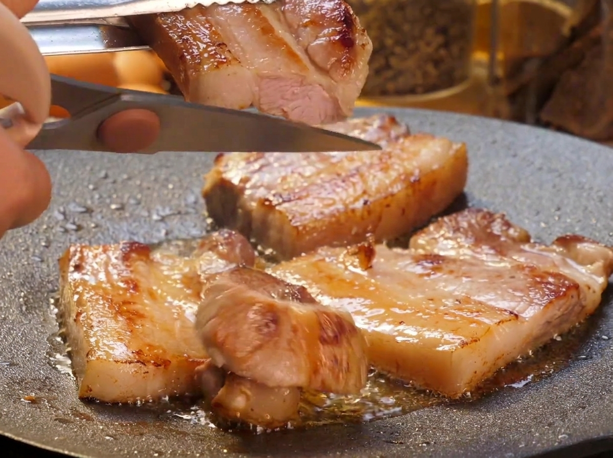 Une main utilisant des ciseaux de cuisine pour couper des tranches épaisses de poitrine de porc saisie en plus petits morceaux au-dessus dune plaque crépitante.