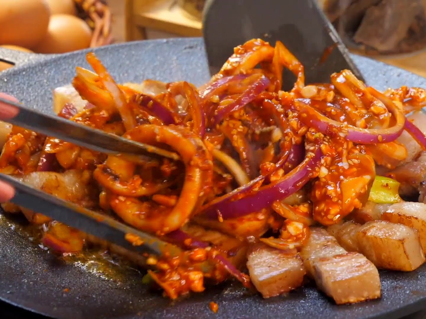 Une spatule et des pinces faisant sauter des anneaux de calmar mariné et des morceaux de poitrine de porc avec des oignons rouges sur une plaque teppanyaki crépitante.