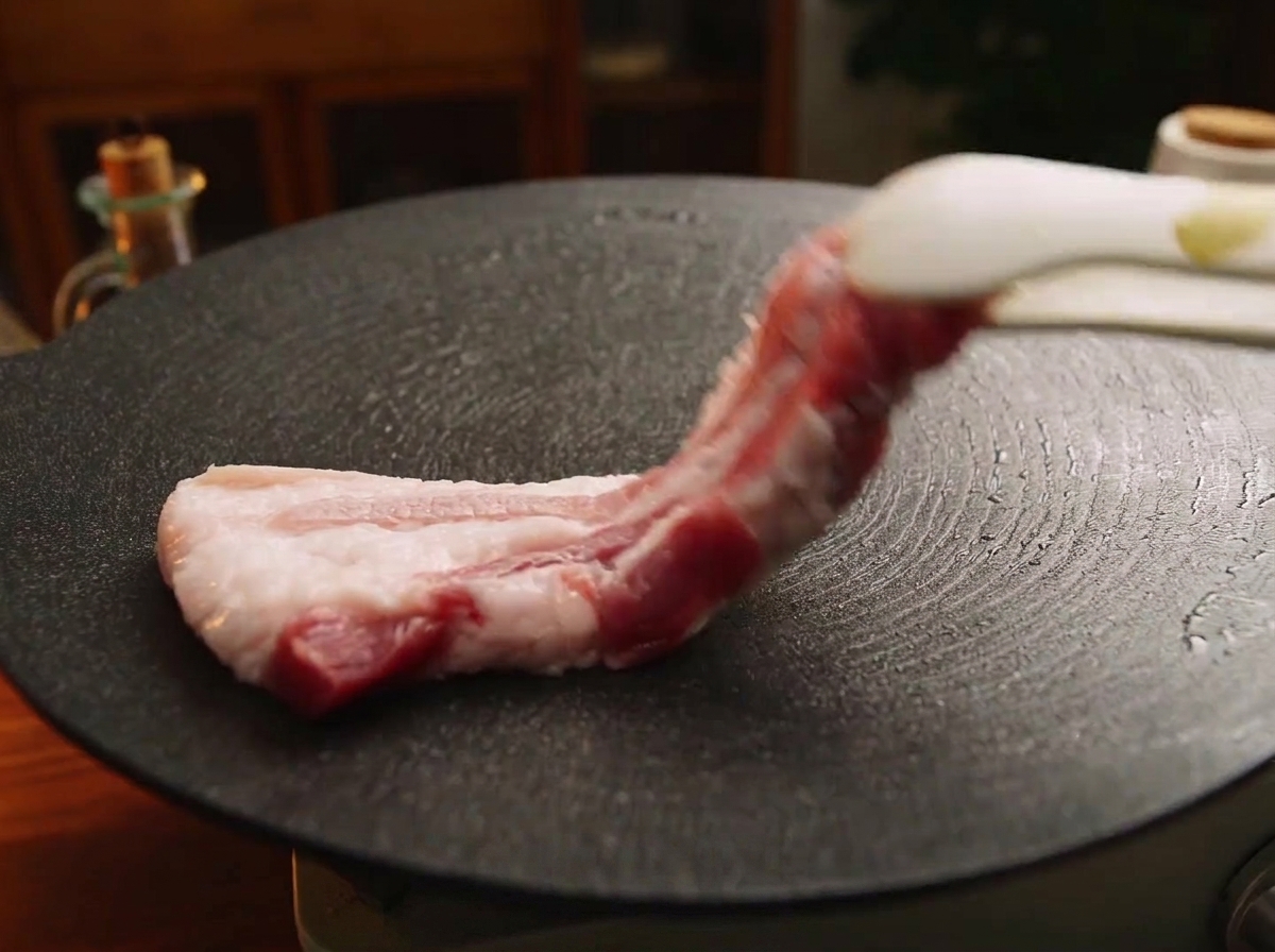 Une pince de cuisine déposant une tranche épaisse de poitrine de porc crue sur une plaque en fonte noire bien chaude.