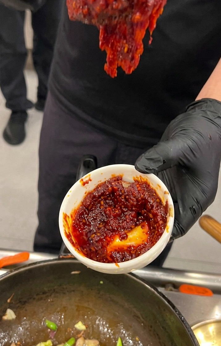 Main gantée de noir tenant un petit bol blanc rempli dune sauce de piment coréenne rouge et épaisse au-dessus du wok.