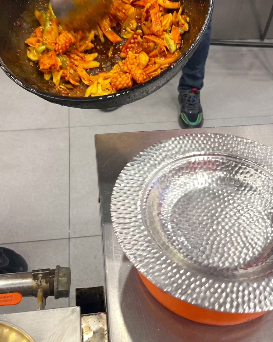 Sauté coréen épicé de calmar et poitrine de porc transvasé depuis un wok vers une assiette de service argentée.