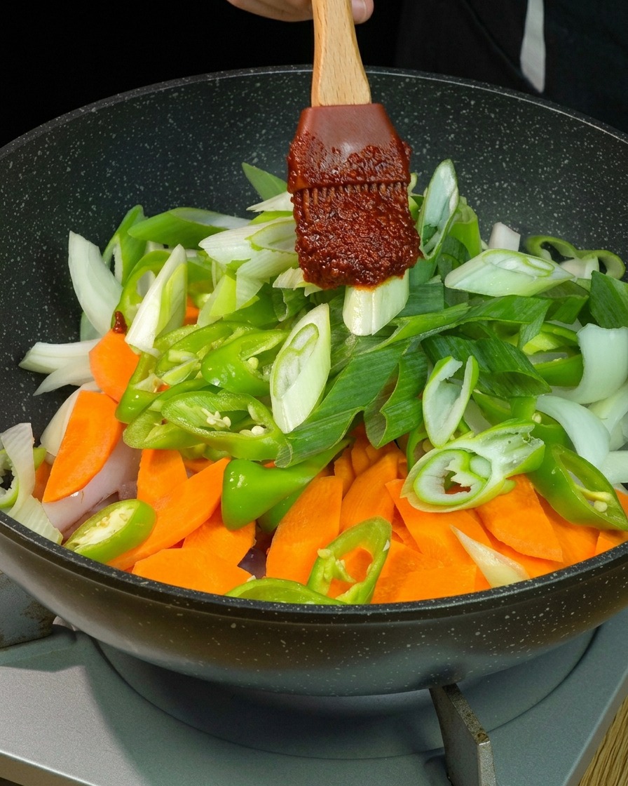 Une cuillérée de sauce au piment rouge déposée sur un mélange coloré de carottes, piments verts, oignons et oignons verts dans une poêle.