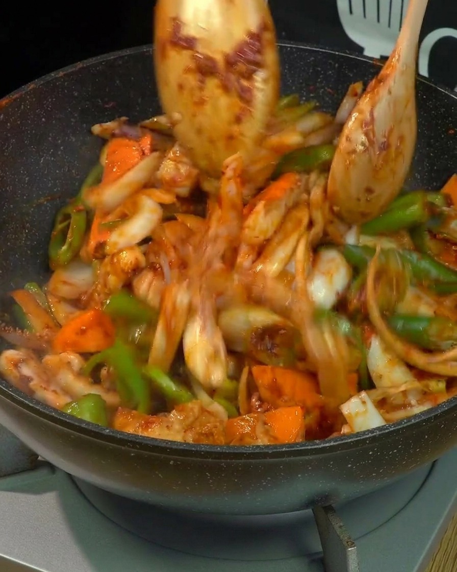 Effet de mouvement montrant de la poitrine de porc, du calamar et des légumes vigoureusement remués avec deux cuillères en bois dans un wok.