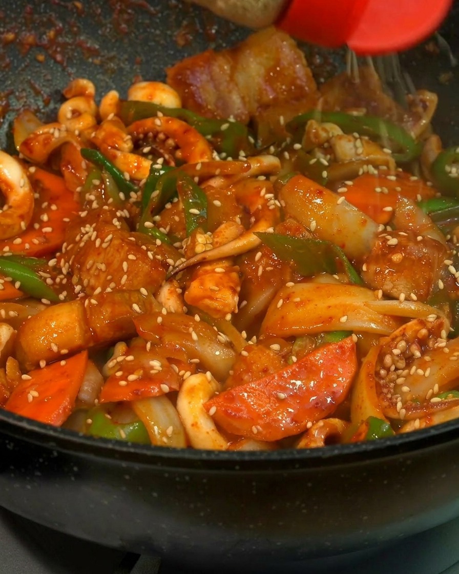 Gros plan sur des graines de sésame blanc saupoudrées sur un sauté brillant de calamar, porc et légumes dans une poêle noire.
