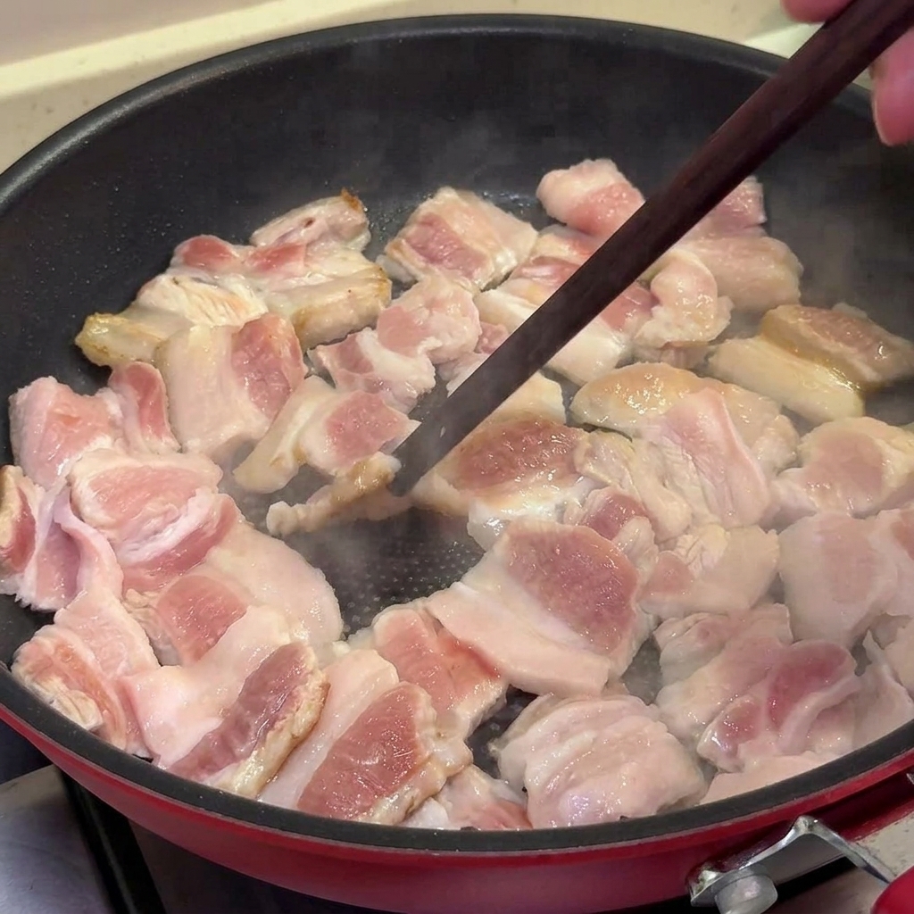 Des tranches de poitrine de porc cuisant dans une poêle antiadhésive, devenant dorées et légèrement grillées, remuées avec des baguettes.