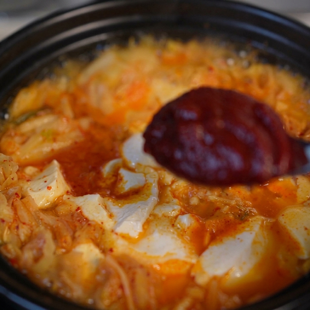 Une cuillère contenant une portion de pâte de piment coréenne rouge foncé et épaisse au-dessus de la soupe frémissante.