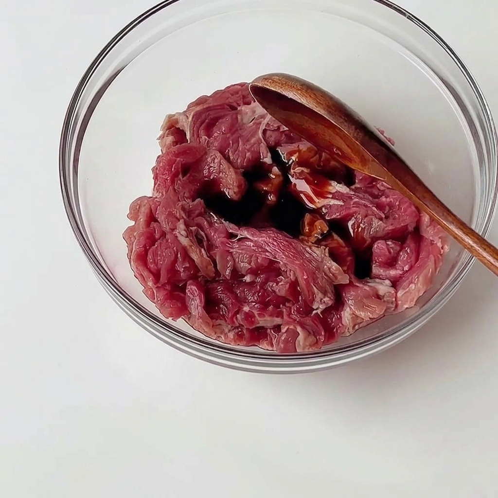 Bœuf cru en fines tranches dans un bol en verre mélangé à des sauces sombres avec une cuillère en bois.
