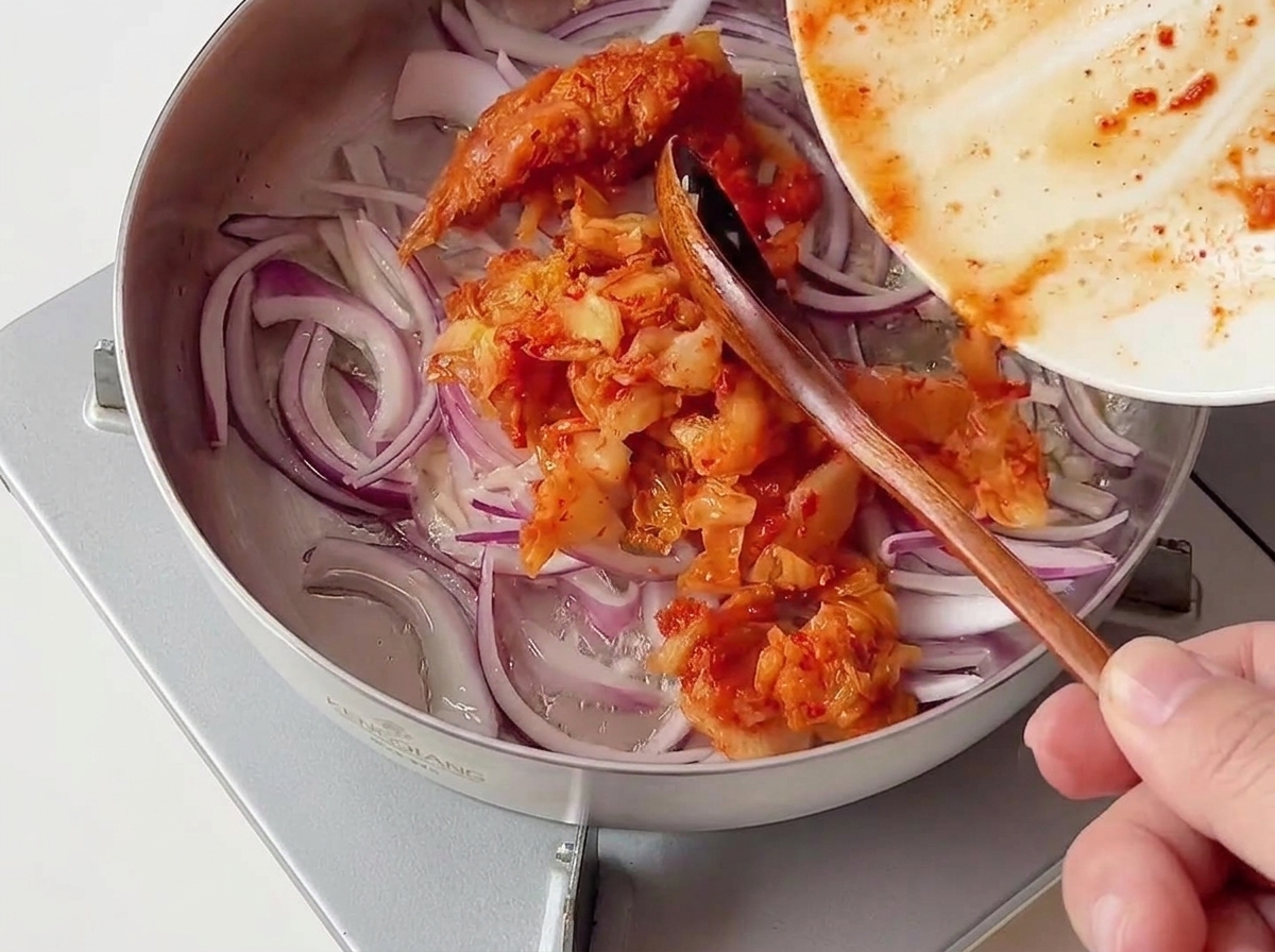 Une portion de kimchi rouge haché ajoutée dans une poêle contenant des oignons rouges émincés.