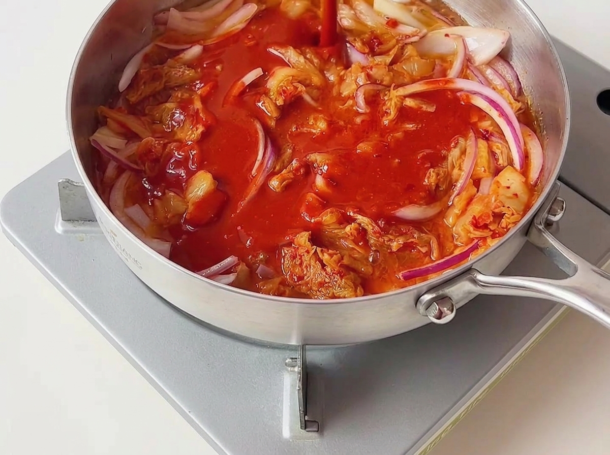Sauce pimentée rouge vif versée sur le kimchi et les oignons rouges sautés dans une poêle argentée.