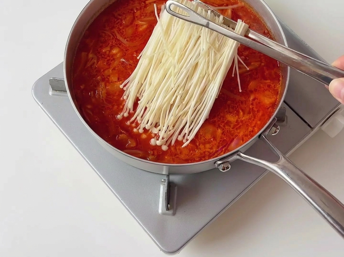 Pince en métal déposant un groupe de champignons enoki blancs dans un ragoût de kimchi rouge bouillonnant.