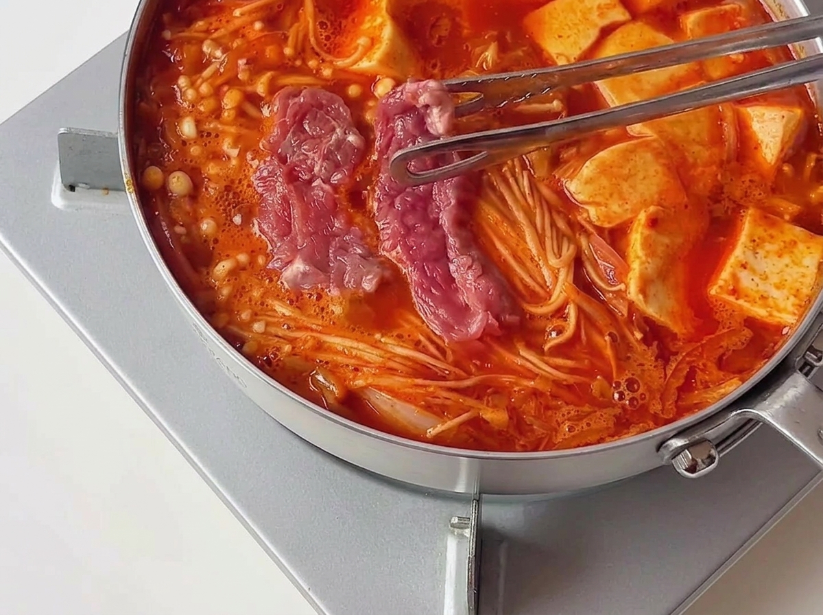 Pinces transférant des morceaux de bœuf mariné cru et rose dans un ragoût chaud et bouillonnant à côté de tranches de tofu.