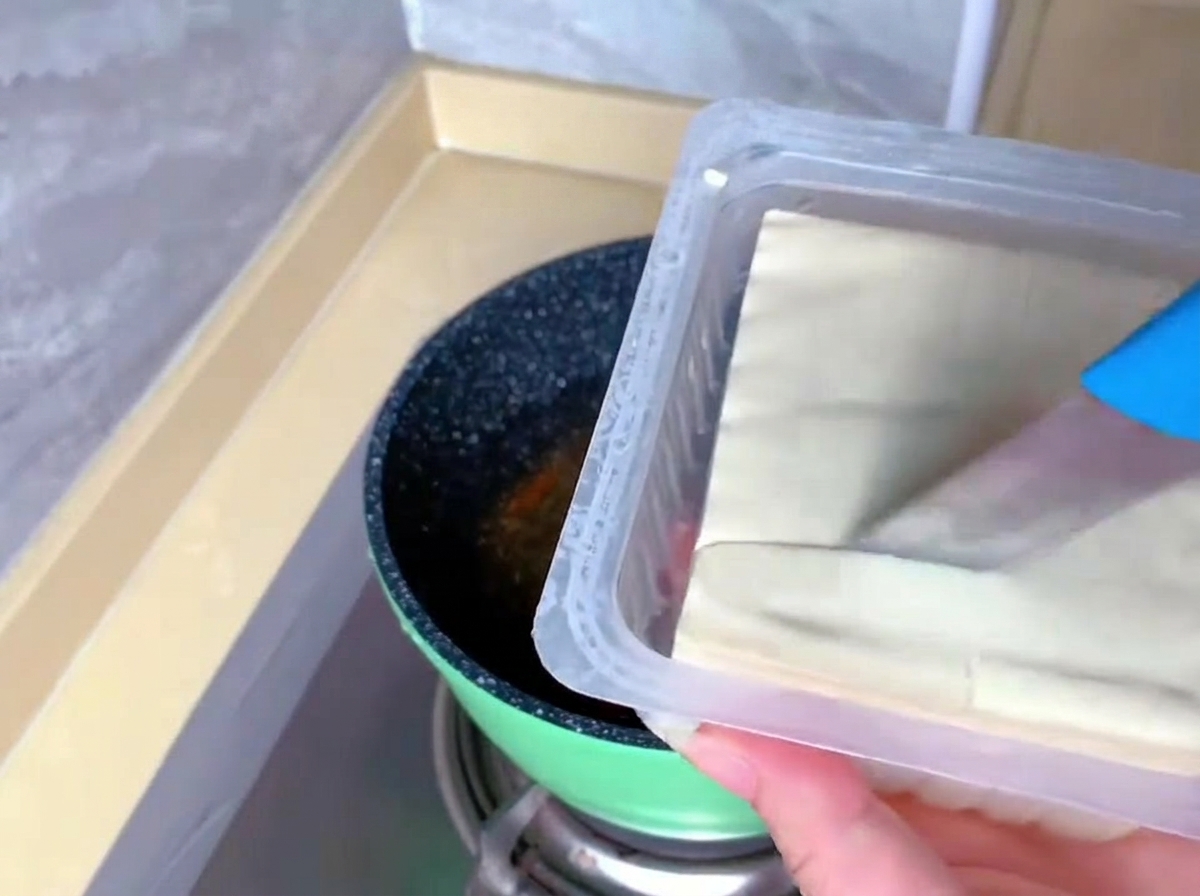 Une personne utilise un couteau bleu pour couper un bloc de tofu soyeux blanc directement dans sa barquette en plastique.