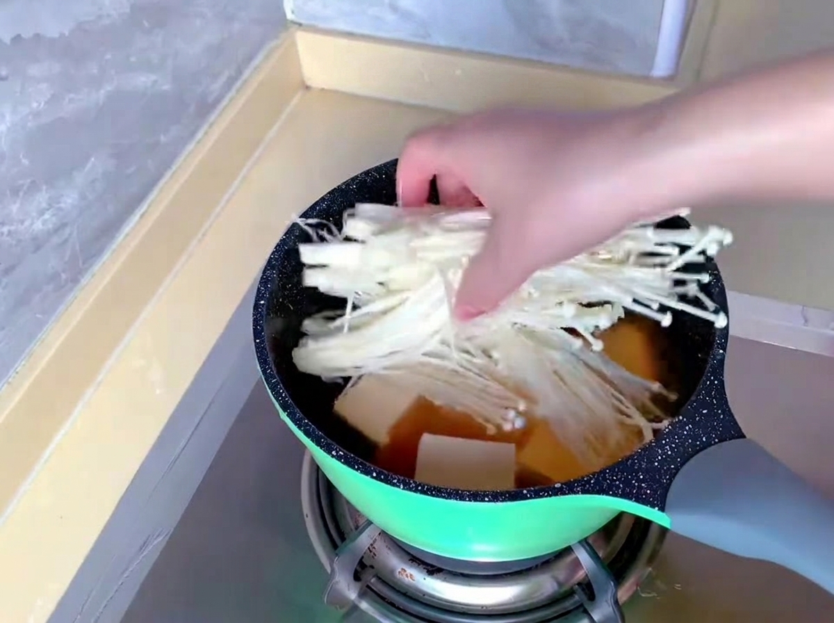 Une main dépose un petit bouquet de longs champignons enoki blancs dans une marmite contenant déjà le bouillon et le tofu.