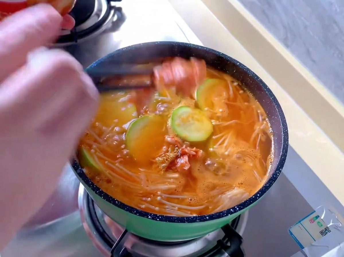 Ajout de morceaux de chou kimchi rouge avec des baguettes dans une petite marmite verte de soupe orange bouillonnante contenant des enoki et des rondelles de courgette.