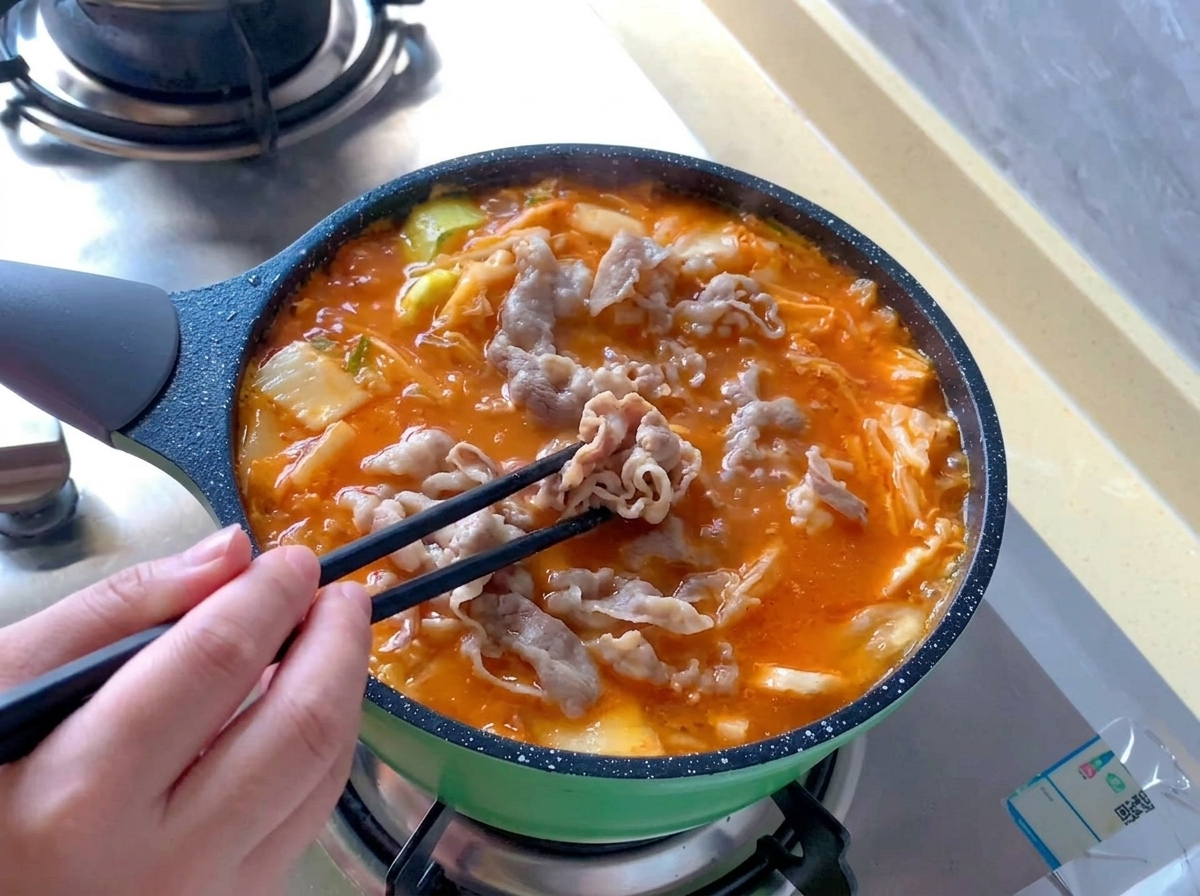 Des roulés de bœuf blanchis sont répartis avec des baguettes noires dans une marmite bouillonnante de soupe au kimchi rouge vif.