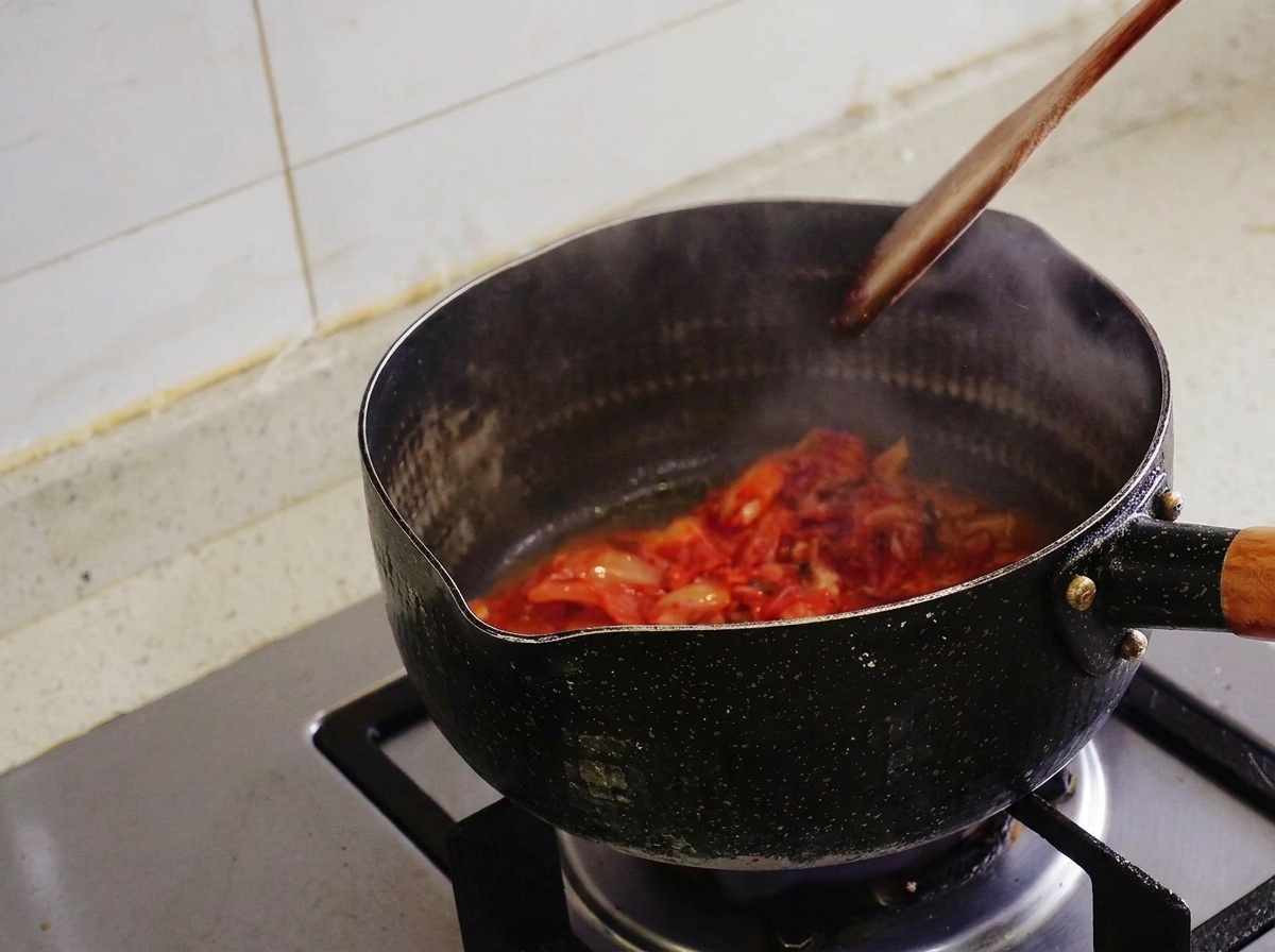 Du kimchi sauté avec une spatule en bois à lintérieur dune casserole noire, doù séchappe de la vapeur.