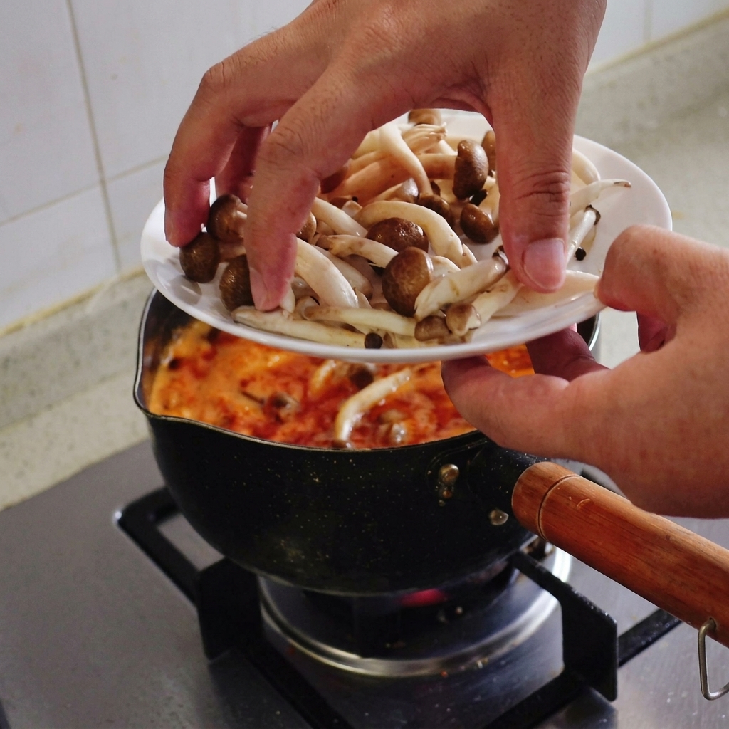 Des mains transférant une poignée de champignons shimeji frais à chapeau brun dune assiette blanche vers une casserole de soupe rouge en ébullition.