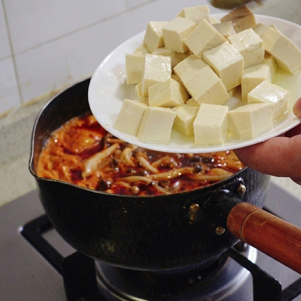 Une main tenant une assiette blanche et y faisant glisser de jolis cubes de tofu soyeux blanc dans un ragoût de kimchi rouge qui mijote.