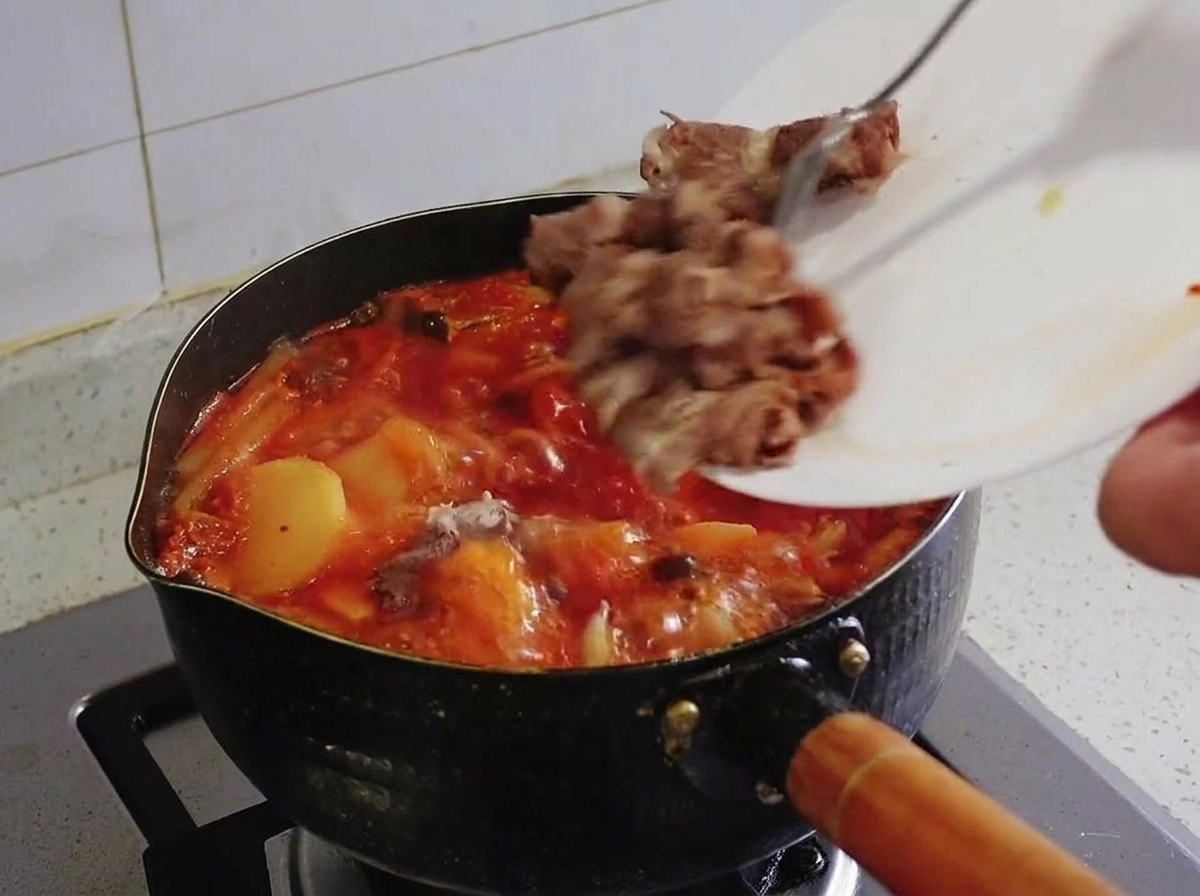 Des morceaux de bœuf gras blanchis glissant dune assiette blanche dans une casserole de ragoût de kimchi rouge en ébullition.