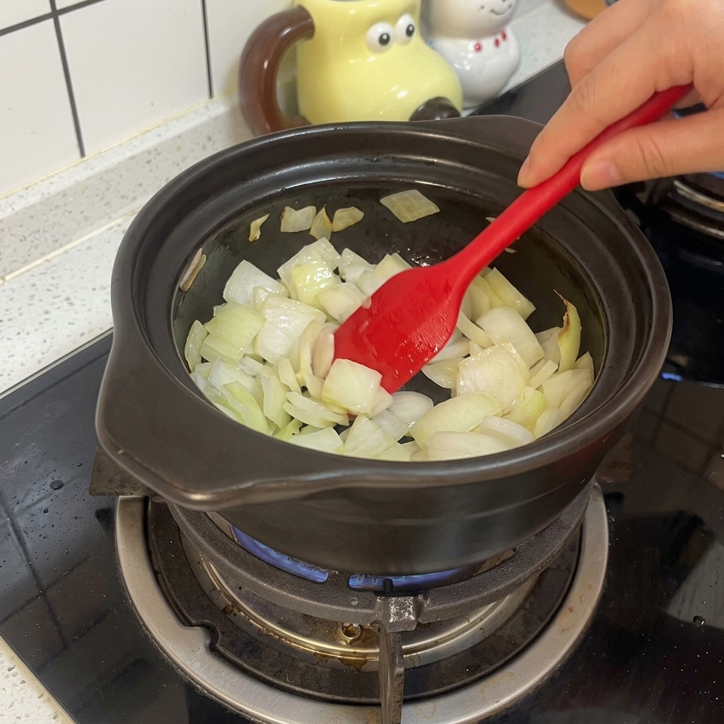 Une spatule en silicone rouge mélange des oignons blancs hachés dans un pot en terre cuite noir sur une cuisinière à gaz.