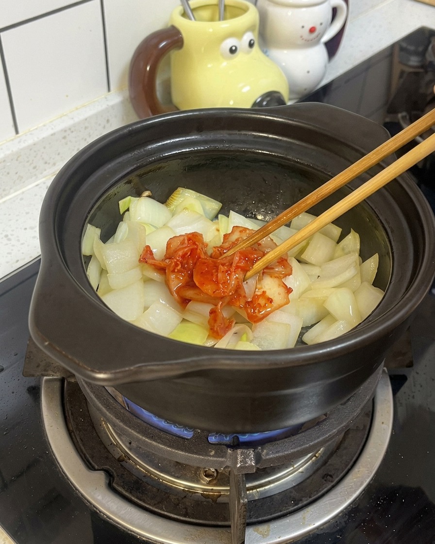 Des baguettes en bois déposent une portion de kimchi rouge fermenté sur des oignons dans un pot en terre cuite noir.