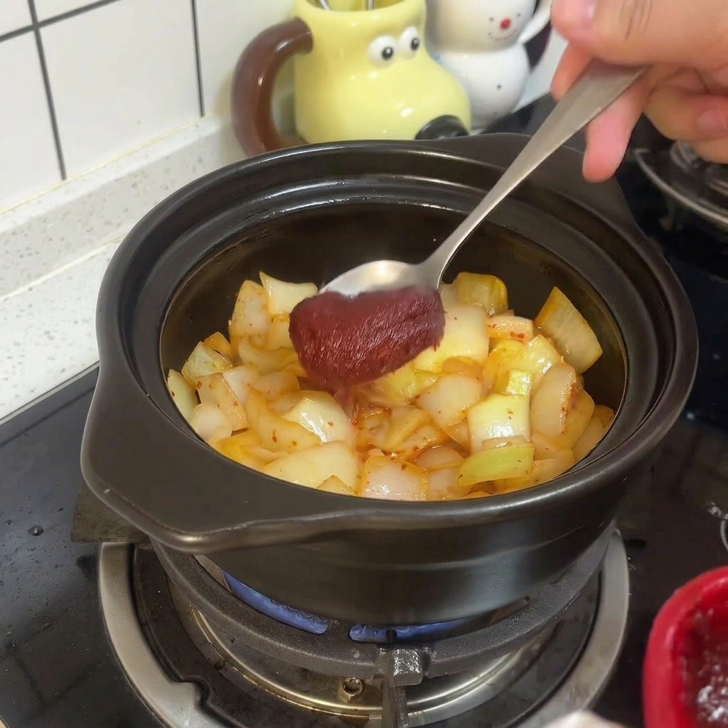 Une cuillère en métal mélange de lépaisse pâte de piment coréenne rouge au kimchi et aux oignons sautés dans le pot en terre cuite.