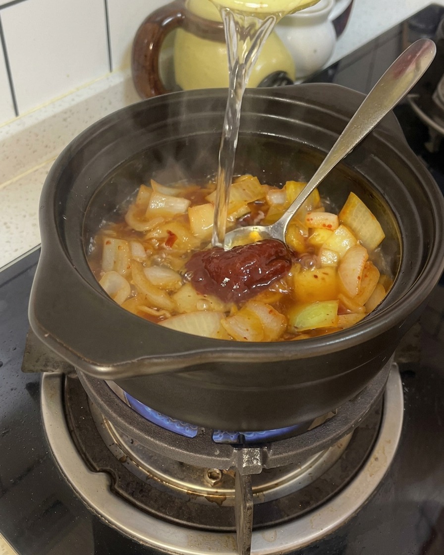 De leau est versée dans un pot en terre cuite noir contenant des oignons assaisonnés et de la pâte de piment rouge.