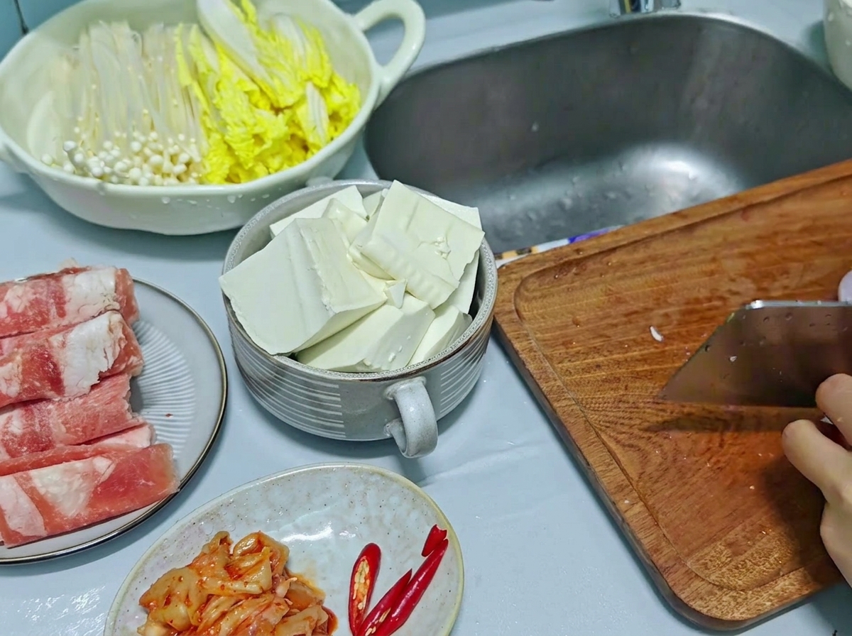 Couperet de cuisine utilisé pour émincer des oignons rouges et des piments rouges sur une planche à découper en bois, avec du tofu tranché visible dans un bol à côté.