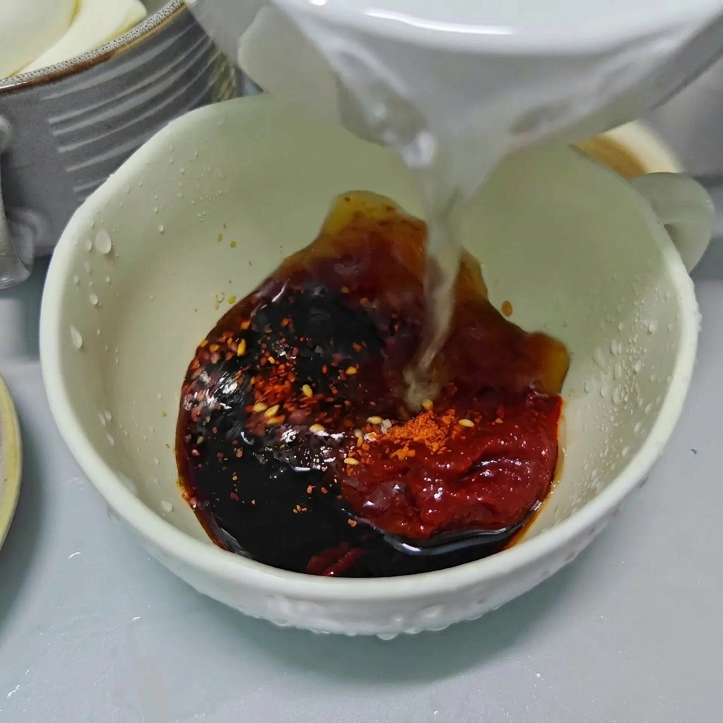 De leau claire versée dans un bol blanc contenant de la sauce soja, de la pâte de piment rouge et des flocons de piment.