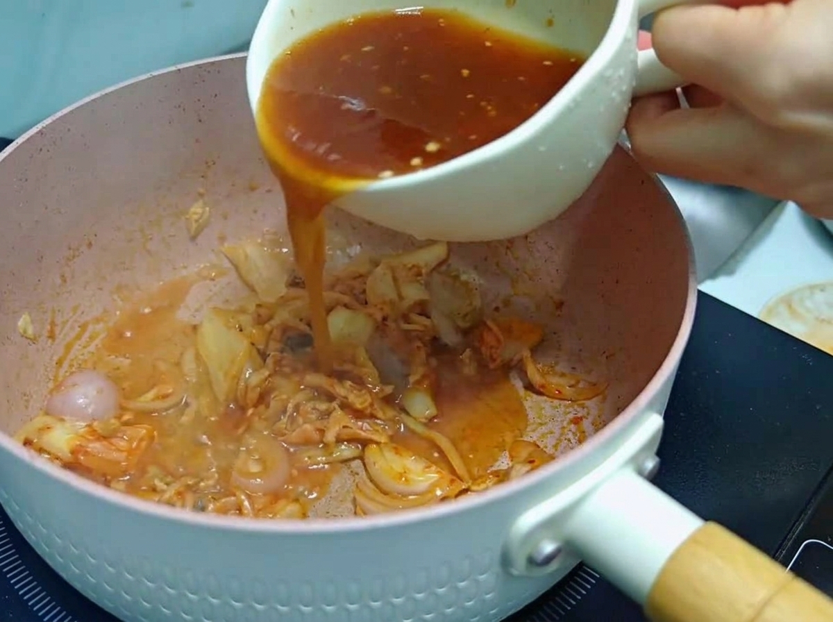 Une personne versant un mélange de sauce rouge-brun dune tasse blanche dans une poêle rose contenant du kimchi et des oignons revenus.